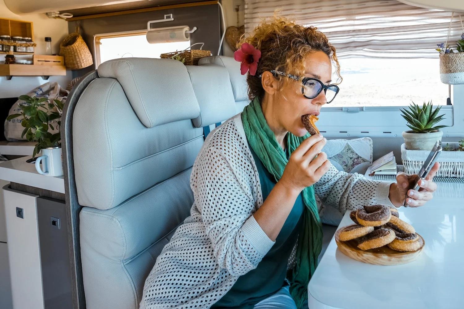 Woman eating donuts while using smartphone in campervan, showing distracted eating habits and automatic food patterns