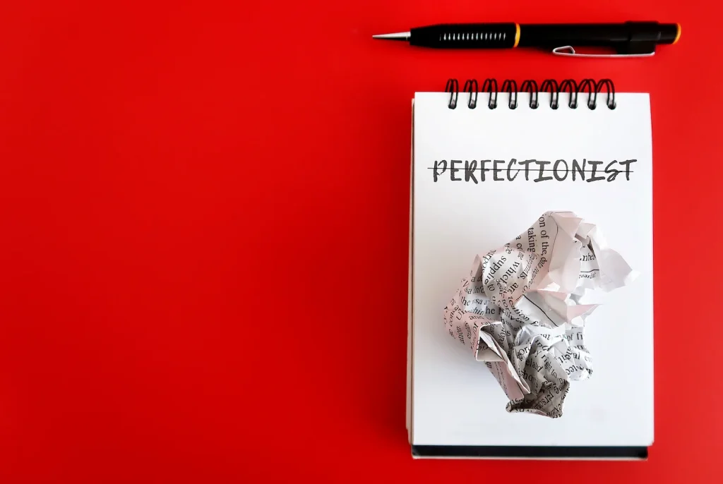 Notebook with the word perfectionist crossed out beside a pen and crumpled paper
